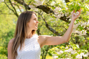 Frau im weißen Kleid betrachtet Blüten an einem Baum. Im Hintergrund ist das frische Grün der Bäume im Frühling zu sehen.