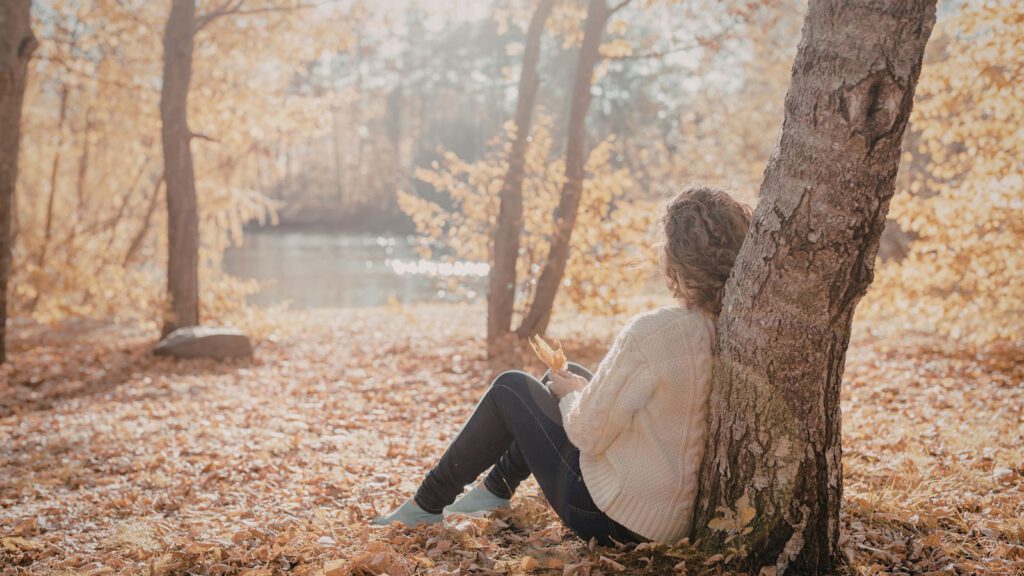 Frau sitzt ruhig an einen Baum gelehnt in einem herbstlichen Wald am Wasser.
